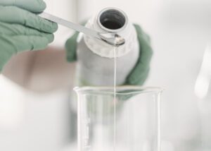 Hands in soft green gloves carefully pouring a clear skincare ingredient in a minimal laboratory setting, representing naturally derived skincare formulation and preservation.