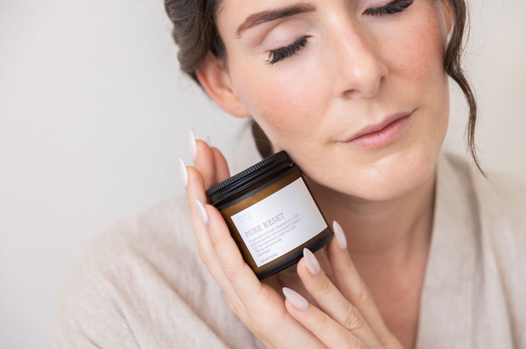 A woman in a linen robe, eyes closed, holding a jar of Indiefog Pure Reset against her cheek